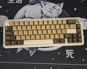 May include: A beige mechanical keyboard with brown and beige keycaps. The keyboard has a silver knob and a white frame. The keyboard is on a black surface with a cat graphic and Japanese characters.