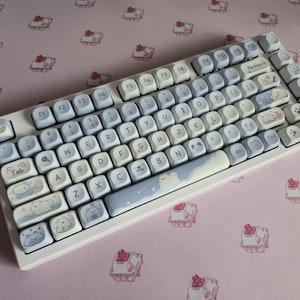 May include: A white mechanical keyboard featuring custom keycaps in shades of light blue and white. The keys display various cartoon animal illustrations, including cats and dogs, alongside standard alphanumeric characters and function keys.