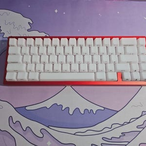 May include: A compact mechanical keyboard with a red frame and white keycaps. It rests on a desk mat with a wave and mountain design in purple, blue, and white. The keyboard has a standard QWERTY layout.