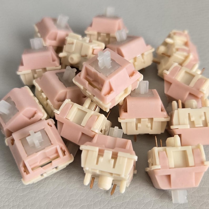 May include: A pile of mechanical keyboard switches in a soft pink and cream color scheme. The switches have a square shape with a clear stem and gold-colored pins. The switches are designed for use in custom keyboards.