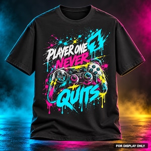 Op de afbeelding: Een zwart t-shirt met korte mouwen en een grafische print van een videogamecontroller met neonroze, blauwe en gele verfspatten. De tekst PLAYER ONE NEVER QUITS staat in een gestileerd lettertype in het midden op de borst gedrukt.