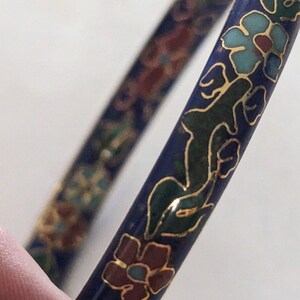 May include: Two blue enamel bangles with floral designs. The bangles feature gold outlines and accents, with flowers in teal, red, and green. The background is white.