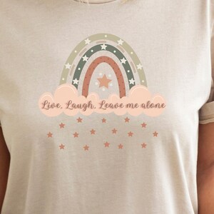 May include: A beige t-shirt with a rainbow graphic and the text "Live. Laugh. Leave me alone". The rainbow is made of green, brown, and orange stripes with white stars. There are small pink stars scattered below the rainbow.