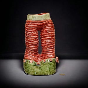 Puede incluir: Escultura de cerámica con forma de pantalón. El pantalón es rojo con estrías horizontales, cintura y puños verdes, y un agujero donde estarían las piernas. Una pequeña moneda de oro está en la superficie.