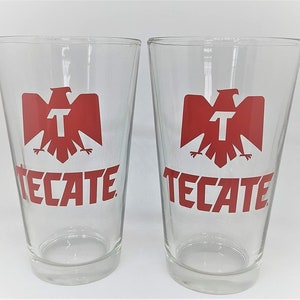 May include: Two clear glass pint glasses with the word "TECATE" in red block letters and a red eagle logo. The glasses are empty and sit side-by-side against a white background.
