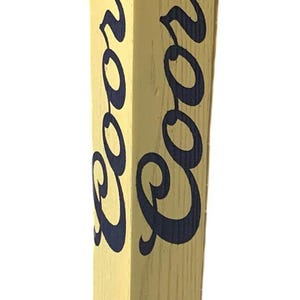 May include: A wooden beer tap handle with the word "Banquet" in red and white at the top. The main body is light yellow with the word "Coors" in navy blue. The bottom has a circular emblem that says "Brewed with Rocky Mountain Water."