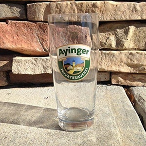 May include: A clear glass beer stein with a green and white logo that says "Ayinger Privatbrauerei".