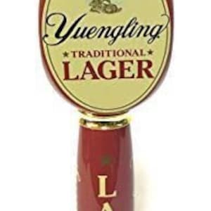 May include: A red and gold beer tap handle with the text "Yuengling Traditional Lager". The handle features a gold eagle emblem at the top and the word "LAGER" in gold lettering. The handle is a classic design.