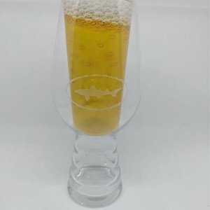 May include: A clear glass beer stein with a shark silhouette etched on the side. The stein is filled with a light-colored beer with a white head.