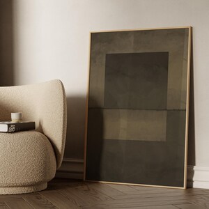 May include: A beige, fluffy armchair with a book and a white cup on top. A large framed abstract painting with a dark brown background and a lighter brown rectangle in the center hangs on the wall behind the chair.