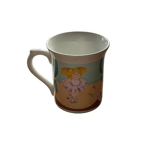 Könnte beinhalten: Ein weißer Keramikbecher mit einer farbenfrohen Illustration einer blonden Figur mit Zöpfen, die ein hellviolettes Tutu und Ballettschuhe trägt. Der Hintergrund zeigt eine Bühne mit türkisfarbenen Vorhängen und einem beigen Boden.