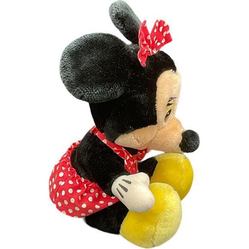 May include: A plush Minnie Mouse doll in a seated position. The doll has black fur, a red and white polka dot skirt, a red bow, and yellow shoes. The doll's face is tan with black eyes and a black nose.