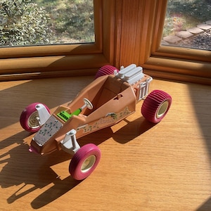 Puede incluir: Un cochecito de dunas Barbie vintage en color beige con ruedas rosas. El coche de juguete tiene un motor blanco y un volante verde. La palabra "Barbie" está impresa en el lateral del coche. El juguete está sobre una superficie de madera.