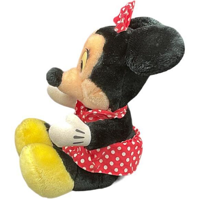 May include: A plush Minnie Mouse doll in a seated position. The doll has a black head and ears, a tan face, and a red bow with white polka dots. She wears a red and white polka dot dress and a matching neckerchief. Her gloves are white, and her shoes are yellow.