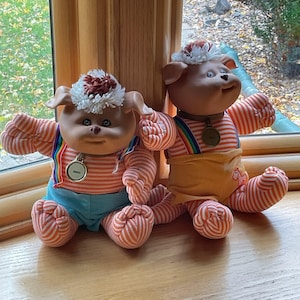 Puede incluir: Dos muñecas vintage estilo Cabbage Patch Kids. Cada muñeca tiene una cara bronceada, orejas caídas y un sombrero blanco y marrón. Llevan camisas a rayas, tirantes arcoíris y pantalones azules o amarillos. Cada muñeca tiene un colgante dorado.