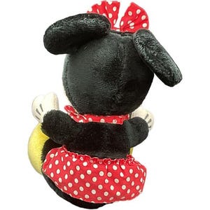 May include: A plush Minnie Mouse doll, viewed from the back, with black fur, large ears, and a red polka-dot bow. She wears a red polka-dot skirt and a matching collar. The doll has white gloves and a yellow object.