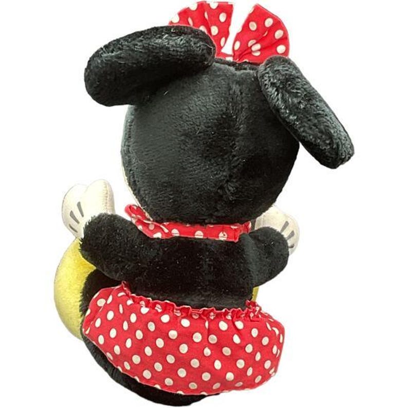 May include: A plush Minnie Mouse doll, viewed from the back, with black fur, large ears, and a red polka-dot bow. She wears a red polka-dot skirt and a matching collar. The doll has white gloves and a yellow object.