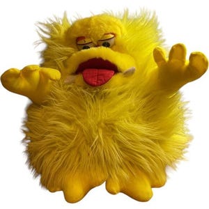 May include: A bright yellow plush monster toy with long, shaggy faux fur, two arms extended outward, and two feet. The creature features a wide mouth with a red tongue, two small white fangs, and two half-closed eyes with orange eyelids.