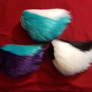 May include: Three faux fur tails in different color combinations. The first tail is teal, white, and teal. The second tail is purple, teal, and purple. The third tail is black, white, and black.