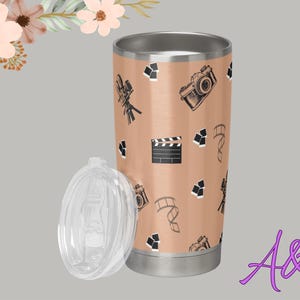 May include: A stainless steel travel mug with a peach-coloured background and a pattern of film cameras, film reels, and clapperboards. The clear lid is off to the side.
