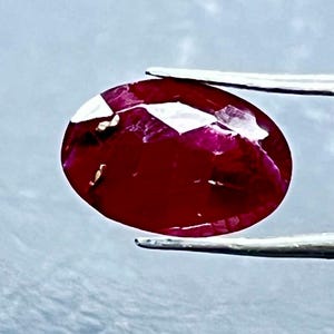 Peut inclure: Une pierre précieuse rouge foncé de taille ovale tenue par une pince en métal argenté. La pierre présente une surface à facettes avec des inclusions internes visibles et des reflets blancs brillants sur le dessus et les bords.