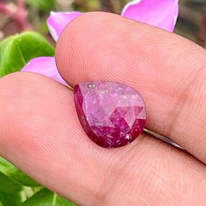 Peut inclure: Une pierre précieuse à facettes en forme de poire, d'une teinte magenta et rose profond. La pierre présente une surface polie avec de multiples facettes triangulaires qui reflètent la lumière, créant un aspect étincelant sur toute sa surface.