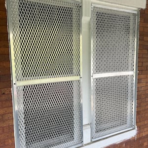 May include: Two windows with silver diamond-patterned security screens. The screens are mounted on a white frame, set against a red brick wall. A white gutter is visible between the windows. The screens appear to be made of metal.