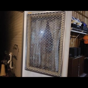 Puede incluir: Un calentador de metal blanco con una rejilla de alambre de malla que cubre el frente. El calentador está montado en una pared y tiene un marco blanco alrededor de la rejilla.