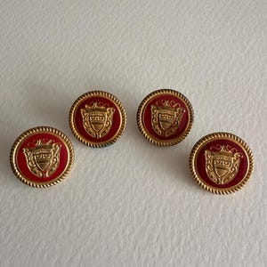 Op de afbeelding: Vier ronde knopen met een rode kern en een goudkleurig embleem. De knopen hebben een goudkleurige, touwachtige rand. Het embleem is een goudkleurig wapenontwerp. De knopen liggen op een wit oppervlak.