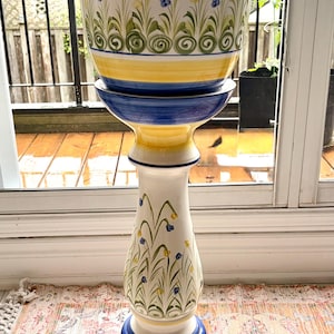 May include: A tall ceramic pedestal planter featuring a cylindrical pot atop a flared base. The piece is decorated with hand-painted blue and yellow bands and a repeating pattern of green stems with small blue and yellow flowers around the exterior.