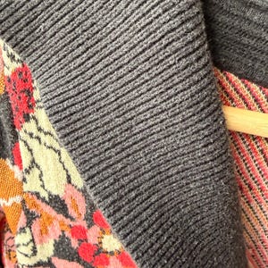 May include: Close-up of a gray knit cardigan with a ribbed texture. The cardigan features a floral patterned panel with pink, orange, and cream-colored flowers and leaves. The garment is hanging on a wooden hanger.