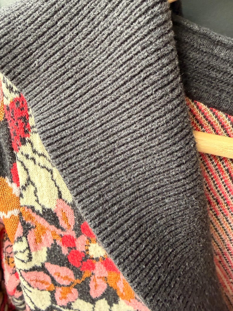 May include: Close-up of a gray knit cardigan with a ribbed texture. The cardigan features a floral patterned panel with pink, orange, and cream-colored flowers and leaves. The garment is hanging on a wooden hanger.