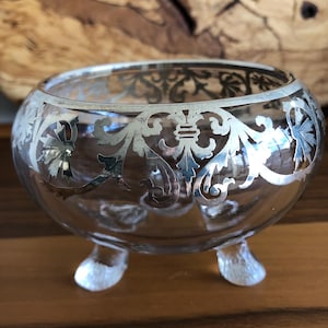 May include: A clear glass bowl with a silver filigree design around the rim and three feet. The bowl is on a wooden surface.