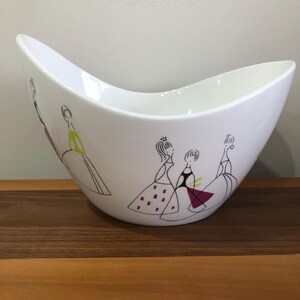May include: White ceramic bowl with a curved shape and a design of four women in black and white line drawings wearing colourful dresses. The bowl is sitting on a wooden surface.
