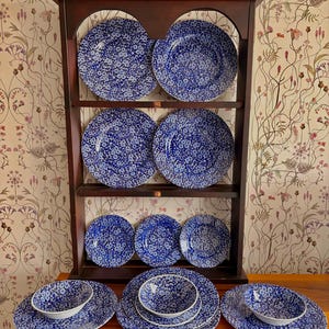 Puede incluir: Una alacena de madera oscura exhibe una colección de vajilla con estampado floral azul y blanco. El juego incluye platos y cuencos de varios tamaños, dispuestos en estantes y una superficie de madera. El estampado floral es un diseño repetitivo de pequeñas flores blancas sobre fondo azul.