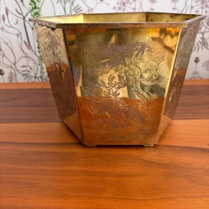 May include: An antique brass planter, octagonal in shape, with etched designs of trees and animals. The brass has a warm, golden colour. The planter is displayed on a wooden surface.