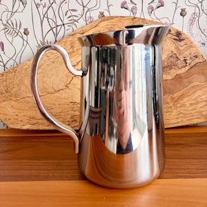 Könnte beinhalten: Eine glänzende Edelstahlkanne mit gebogenem Griff. Die Kanne hat eine breite Öffnung und einen abgerundeten Boden. Der Griff ist an der Seite befestigt. Die Kanne steht auf einer Holzoberfläche.