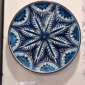 May include: A round, decorative plate with a hand-painted floral design in shades of blue and white. The plate features a central starburst pattern surrounded by stylized floral motifs. The rim is a darker blue, and the plate is mounted on a wall.