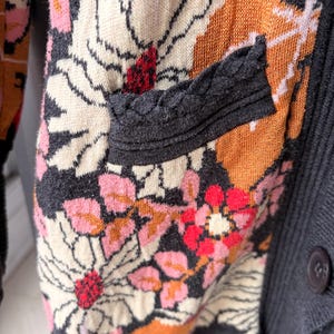May include: Close-up of a floral patterned cardigan. The sweater features a mix of colors, including pink, orange, cream, and red, set against a dark gray background. The design includes a pocket with a braided trim and a button closure.