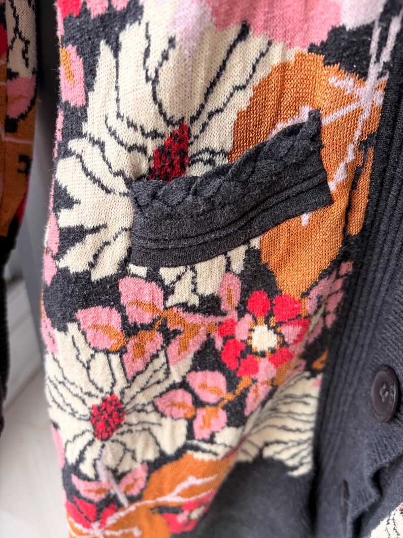 May include: Close-up of a floral patterned cardigan. The sweater features a mix of colors, including pink, orange, cream, and red, set against a dark gray background. The design includes a pocket with a braided trim and a button closure.