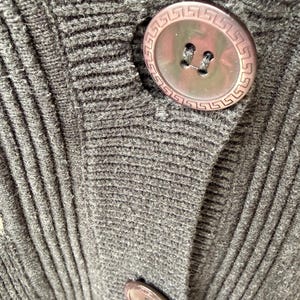 May include: Close-up of a dark gray knit cardigan with large, round, brown buttons. The buttons have a decorative Greek key border. The knit fabric has a ribbed texture.