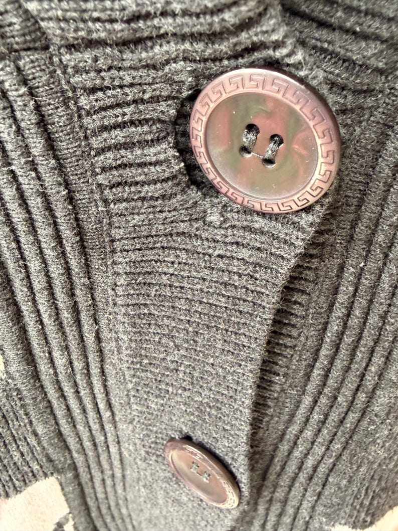 May include: Close-up of a dark gray knit cardigan with large, round, brown buttons. The buttons have a decorative Greek key border. The knit fabric has a ribbed texture.