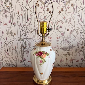 May include: White ceramic table lamp with a gold base and accents. The lamp features a floral design with red, pink, and yellow roses. The lamp has a brass neck and a brown finial. The lamp is approximately 18 inches tall.
