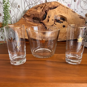 May include: Three clear glass drinking glasses with gold rims and a gold logo. The glasses are arranged on a wooden surface. The logo appears to be a crown with the word "DOW" underneath. The background includes a wooden slab and floral wallpaper.