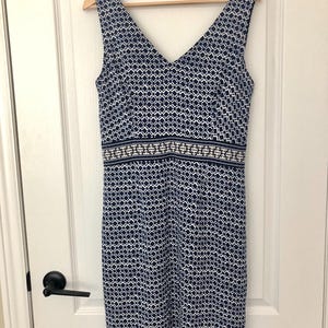 May include: Sleeveless V-neck dress with a geometric pattern in navy and white. The dress has a contrasting band at the waist and is hanging on a wooden hanger. The dress is made of a lightweight fabric.