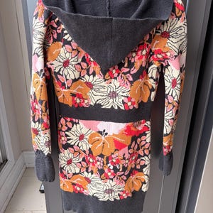May include: A long-sleeved hooded sweater with a floral pattern in shades of orange, pink, and cream on a black background. The hood, cuffs, and hem are a solid dark gray. The sweater is hanging on a wooden hanger.
