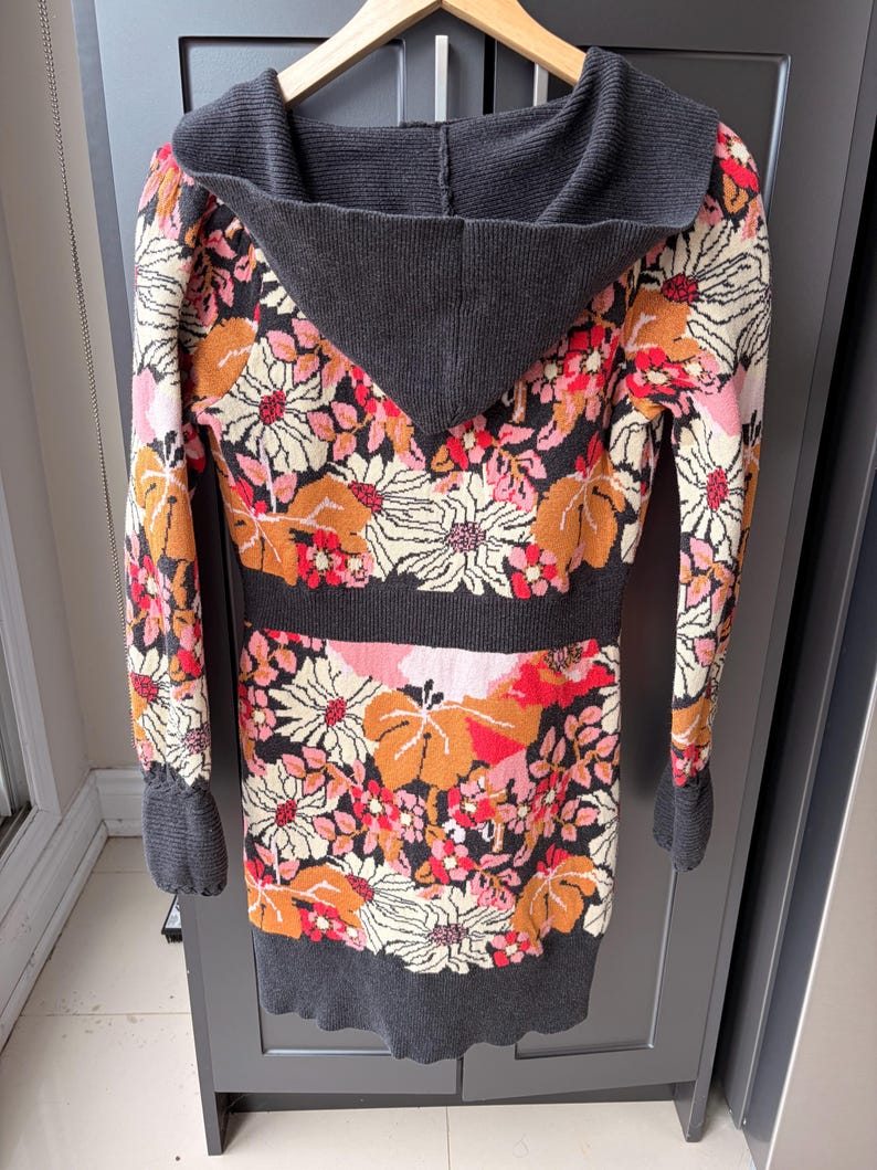 May include: A long-sleeved hooded sweater with a floral pattern in shades of orange, pink, and cream on a black background. The hood, cuffs, and hem are a solid dark gray. The sweater is hanging on a wooden hanger.