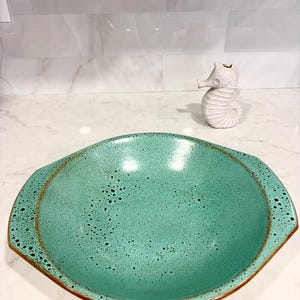 May include: A large, turquoise ceramic serving bowl with a speckled finish and a brown rim. The bowl has a unique, slightly oval shape with integrated handles. A small, white seahorse figurine is in the background.