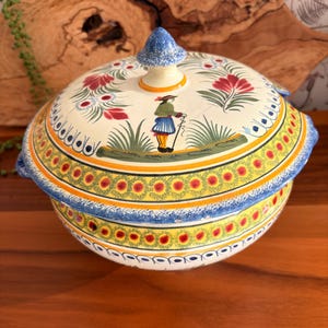 Peut inclure: Une soupière ronde en céramique avec un couvercle orné d'un motif d'art populaire peint à la main. La surface crème présente des motifs floraux rouges, du feuillage vert et un personnage central en jupe bleue. Des bandes jaunes et bleues décorent.