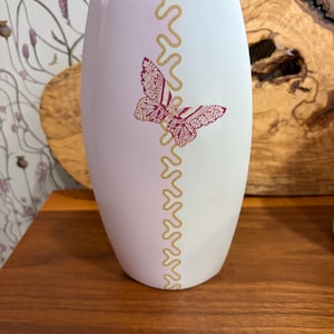 May include: White ceramic vase with a butterfly design. The vase features a pink butterfly and a gold wavy line pattern. The vase is on a wooden surface.